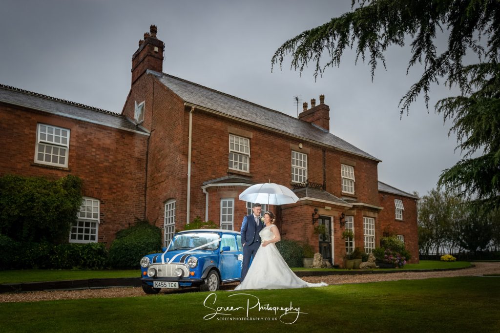 Married couple in front of Swancar Farm Country House in rain with umbrella and classic mini car