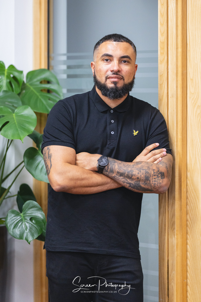 commercial photography nottingham business corporate headshot of a man leaning against a glazed wall with arms folded showing the waist upwards and plant in background